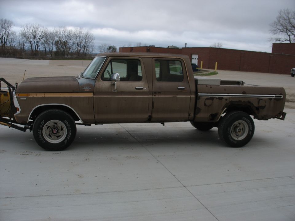 Brown 1970s Ford crew cab pickup with front snow plow and rusty rear fender, parked on wet pavement