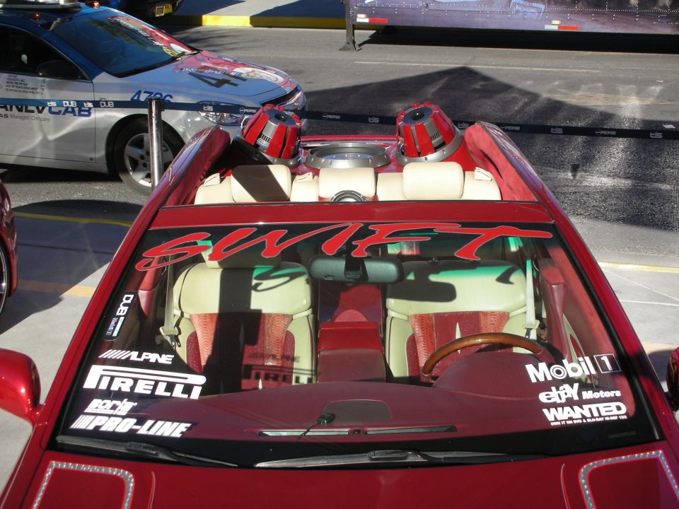 Red convertible with 'SWIFT' on windshield, custom interior, and two large red speakers in the back. A silver car with 'DUB' logos is parked nearby