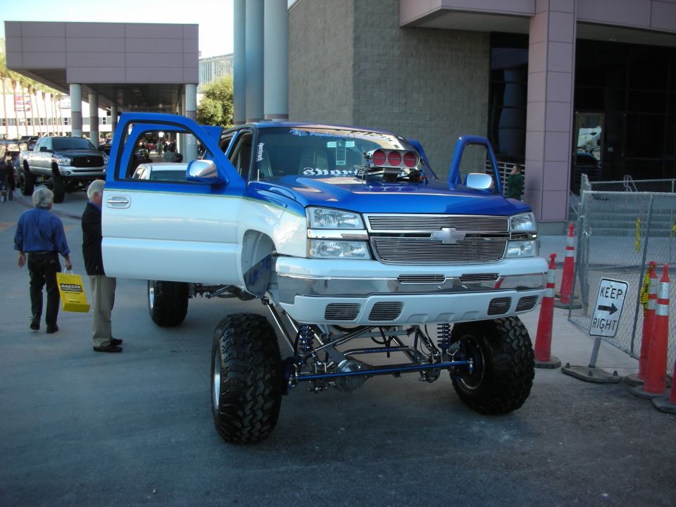 Blue and white lifted pickup truck with open doors and exposed engine components. Another pickup truck is parked behind it