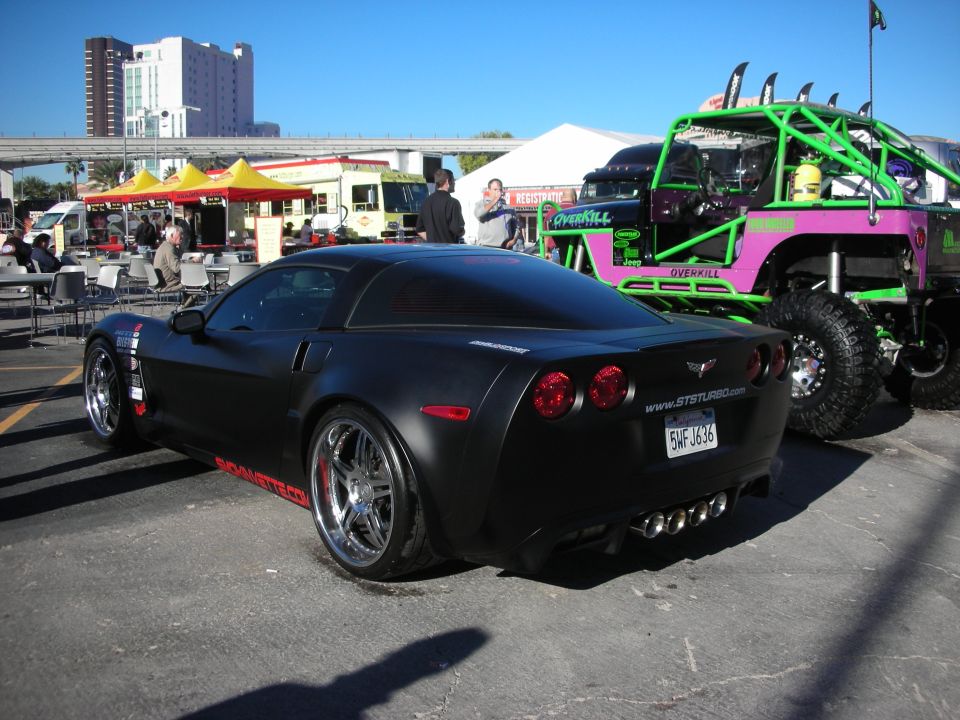 Black Chevrolet Corvette with red accents and quad exhaust pipes, parked next to a custom off-road vehicle