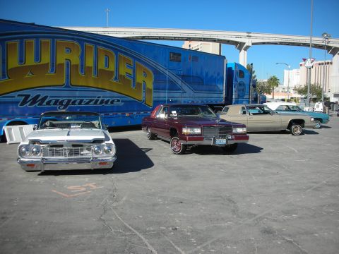 1998 Ford Crown Victoria - Three lowrider cars are parked in front of a Lowrider Magazine trailer on a sunny day.