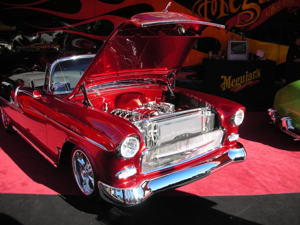 Red 1955 Chevrolet convertible with engine exposed, parked on red carpet. Another car is visible to the right