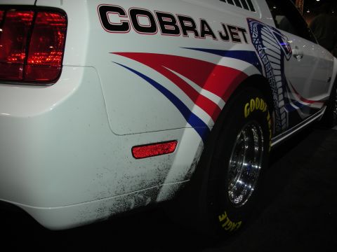 1998 Ford Crown Victoria - A white Ford Mustang displays 'Cobra Jet' graphics and racing tires, seen from the rear quarter panel.