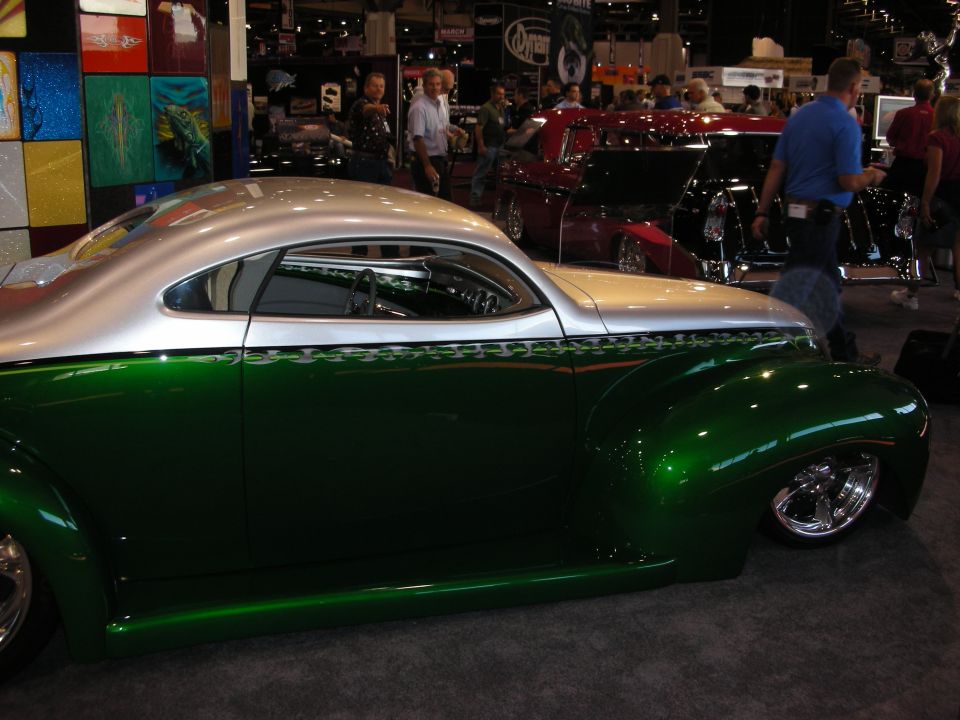 Green and silver custom hot rod coupe with flame pinstriping, seen at a car show with other vehicles in the background