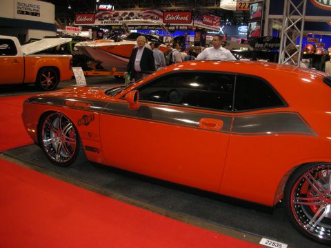 1998 Ford Crown Victoria - An orange Dodge Challenger is displayed at a car show with other vehicles and booths in the background.