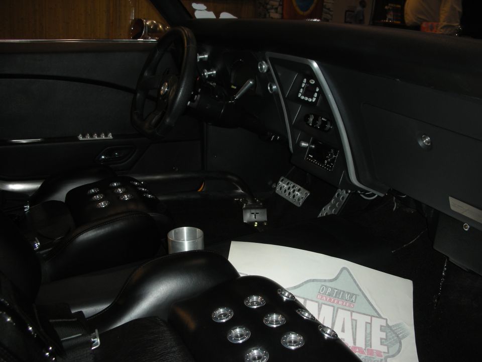 Car interior with bucket seats, custom steering wheel, and dashboard with digital gauges. A paper with 'OPTIMA' logo is on the floor