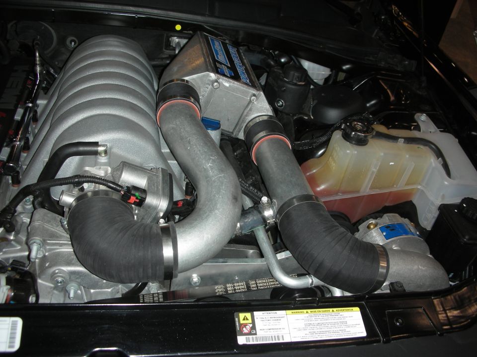 Engine bay with a large silver supercharger, black hoses, and a coolant reservoir