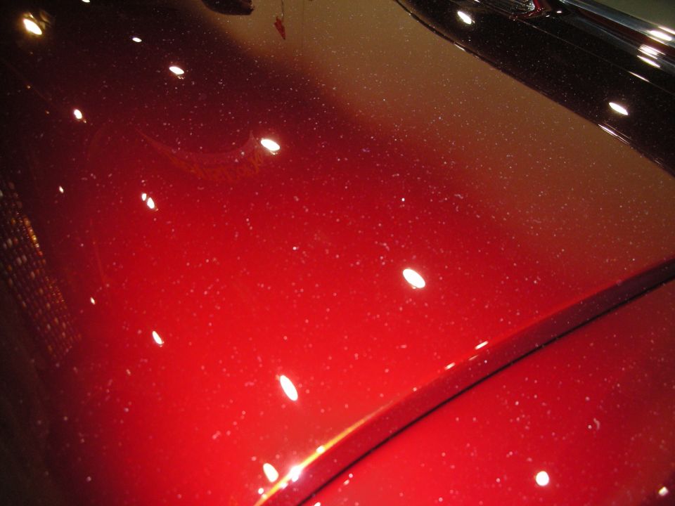 Close-up of a red car hood covered in dust specks, with reflections from overhead lights
