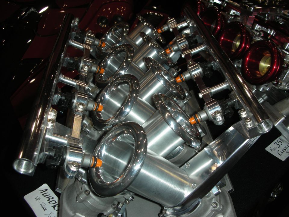 Close-up of a V8 engine intake manifold with chrome trumpets and fuel injectors. Text on a tag reads 'AURORA V8 Race X'
