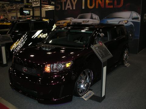 1998 Ford Crown Victoria - Customized dark maroon Scion xB with chrome wheels displayed under a 'Stay Tuned' banner at an indoor auto show.
