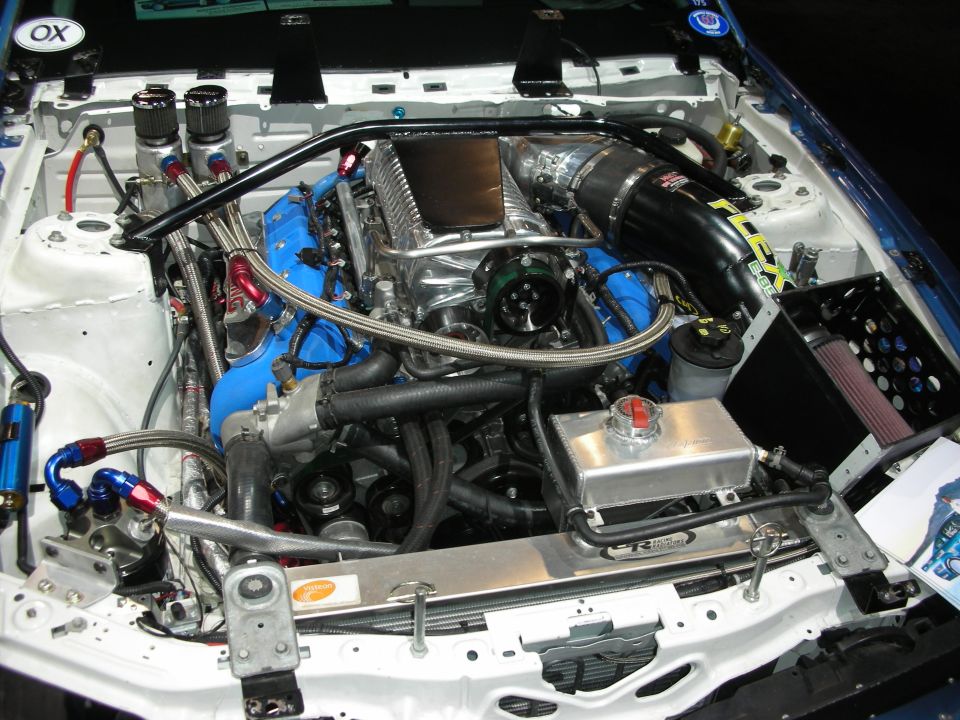 Engine bay with supercharger, blue intake manifolds, and custom piping. Some text visible on air intake