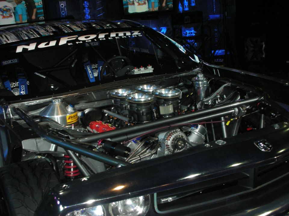 Engine bay of a dark car with 'NaFormz Engineering' on the windshield. Features multiple carburetors and a roll cage