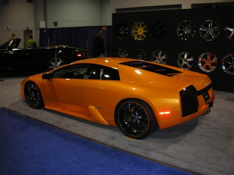 1998 Ford Crown Victoria - An orange sports car is displayed indoors next to a wall of custom rims, aftermarket rims.
