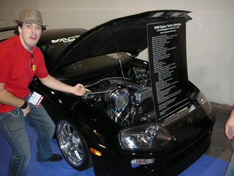 1998 Ford Crown Victoria - A man points to the engine of a black 1995 Toyota Supra Turbo Hardtop displayed with its hood open.
