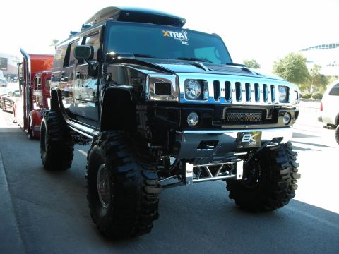 1998 Ford Crown Victoria - A heavily modified Hummer H2 with oversized tires and a roof-mounted cargo box is parked beside a semi-truck.