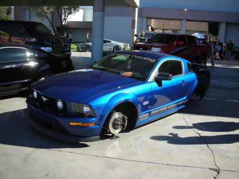 1998 Ford Crown Victoria - Blue 2005 Ford Mustang stripped of its wheels, parked at an outdoor event with other vehicles present.