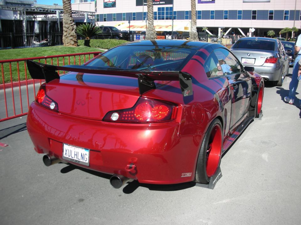 Red Infiniti coupe with large rear spoiler and custom wheels. License plate reads XULHLNG. Another car is parked behind it
