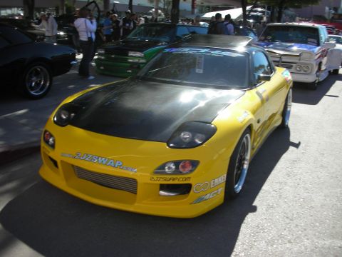 1998 Ford Crown Victoria - A modified yellow Mazda RX-7 is parked at an outdoor event with other vehicles and spectators.