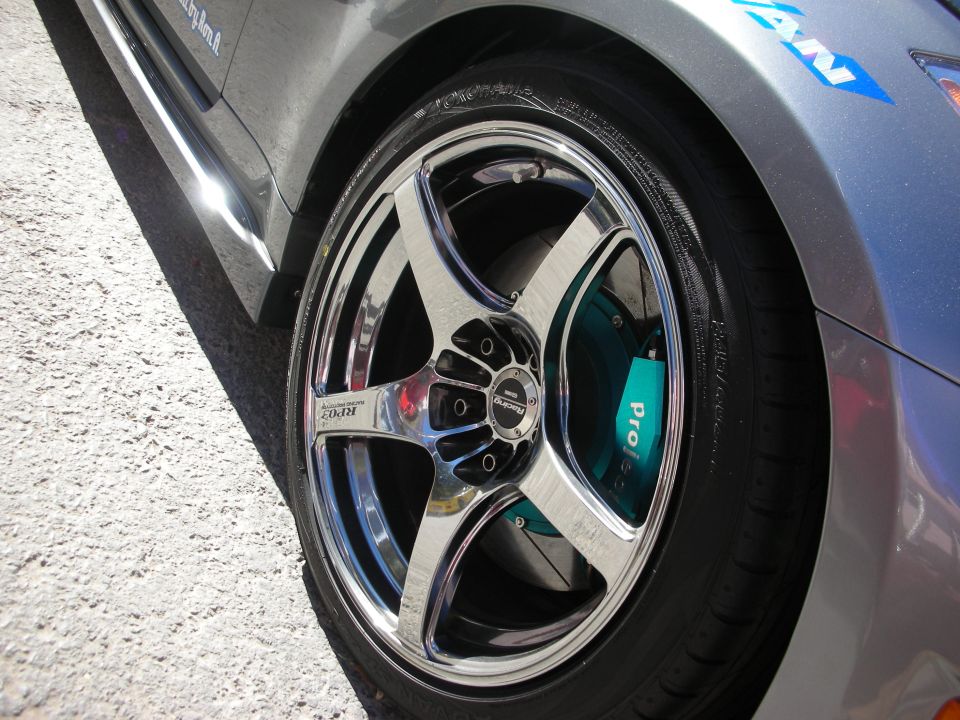 Chrome five-spoke wheel with teal "Project" brake caliper on a gray car