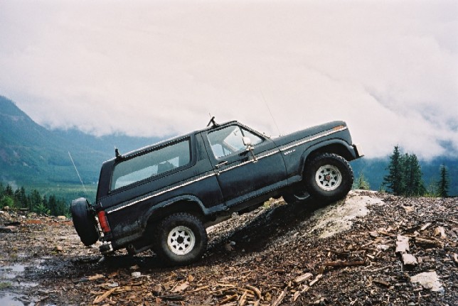 Dark Ford Bronco SUV climbing a steep dirt slope, side view, with foggy mountains behind