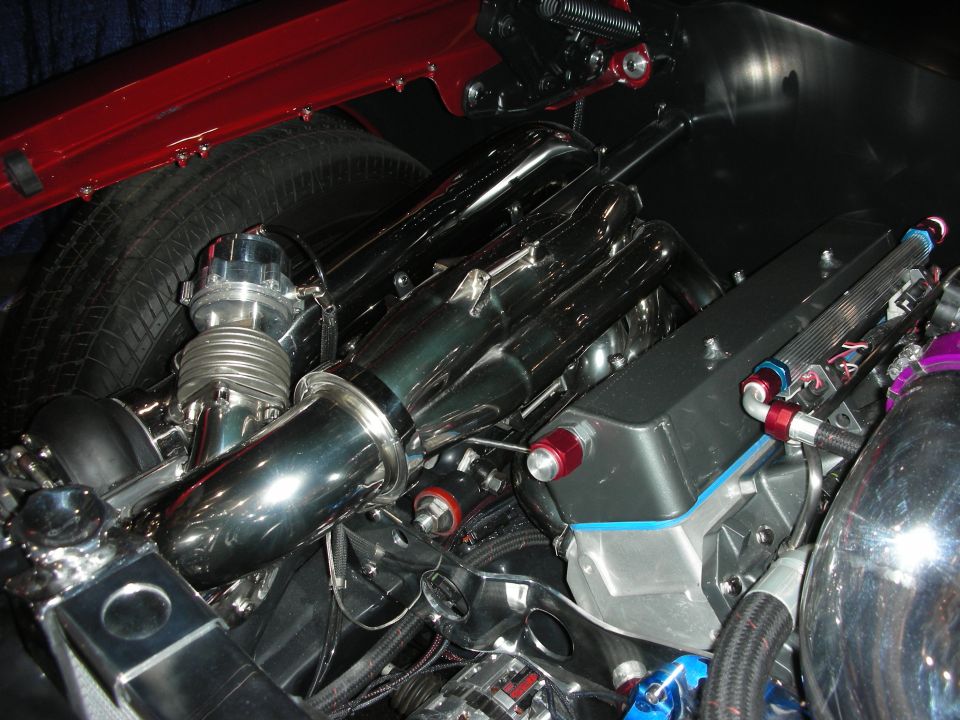 Close-up of a shiny engine bay with a turbocharger, intercooler, and custom plumbing. Red fender liner visible above a tire