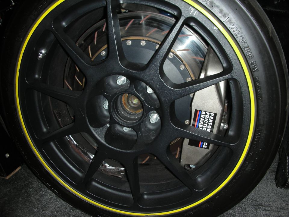 Close-up of a black multi-spoke wheel with a yellow stripe on the tire, showing a large brake caliper labeled 'PERFORMANCE FRICTION BRAKE'