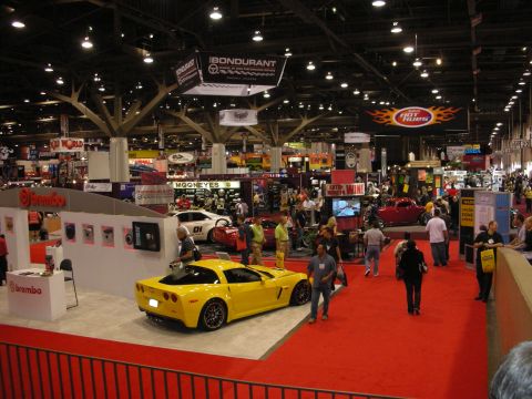 1998 Ford Crown Victoria - A bright yellow sports car is showcased at an indoor automotive event, surrounded by exhibition booths and attendees.