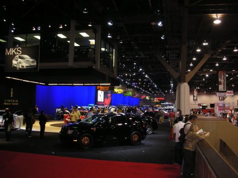 1998 Ford Crown Victoria - A black Lincoln MKS is showcased at an automotive exhibition, with various displays and attendees around.