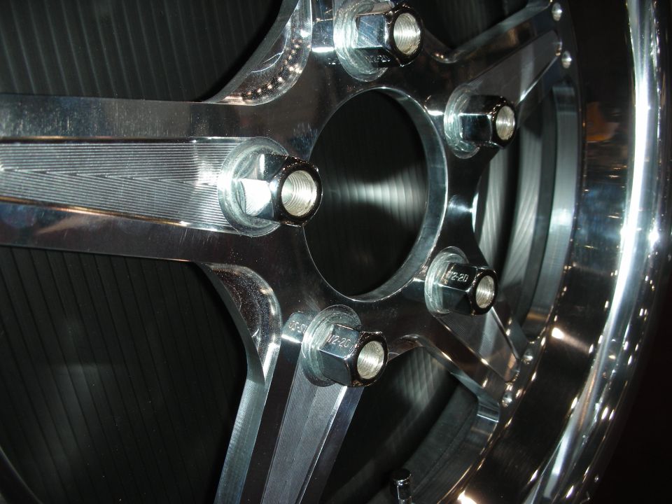 Close-up of a shiny chrome wheel with five lug nuts. Some text is visible on the lug nuts