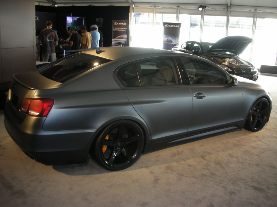 Matte gray sedan with black wheels and orange brake calipers. Another car with its hood open is visible behind it