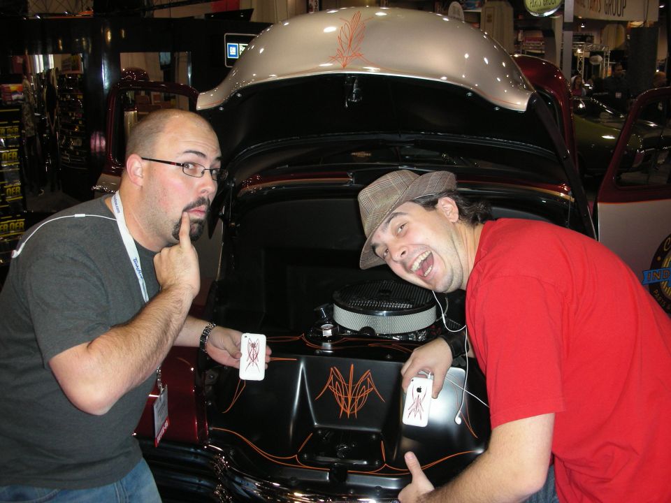 Two men pose with phones in front of a car's engine bay, which has custom pinstriping