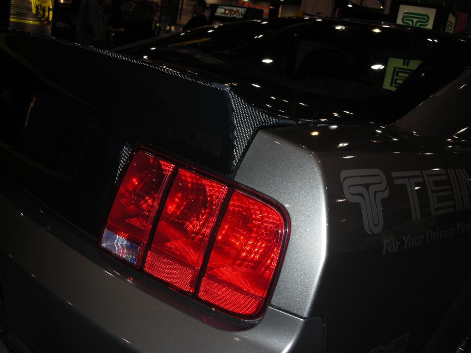 Gray car rear with carbon fiber spoiler and triple-bar taillight. Text "TEIN For Your Driving Pleasure" visible