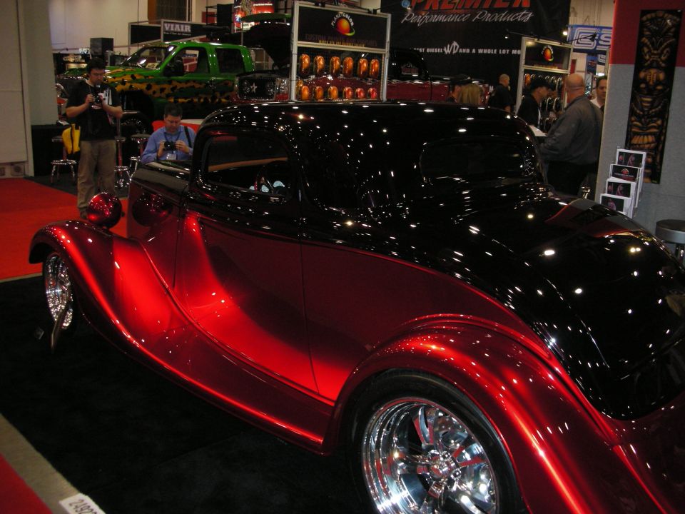 Two-tone red and black hot rod coupe with custom wheels at a car show. Green pickup truck in background