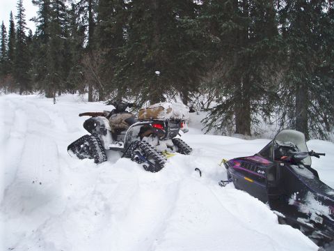 2000 Ford E350 - An ATV equipped with tracks is stuck in deep snow in a wooded area, with a snowmobile nearby.