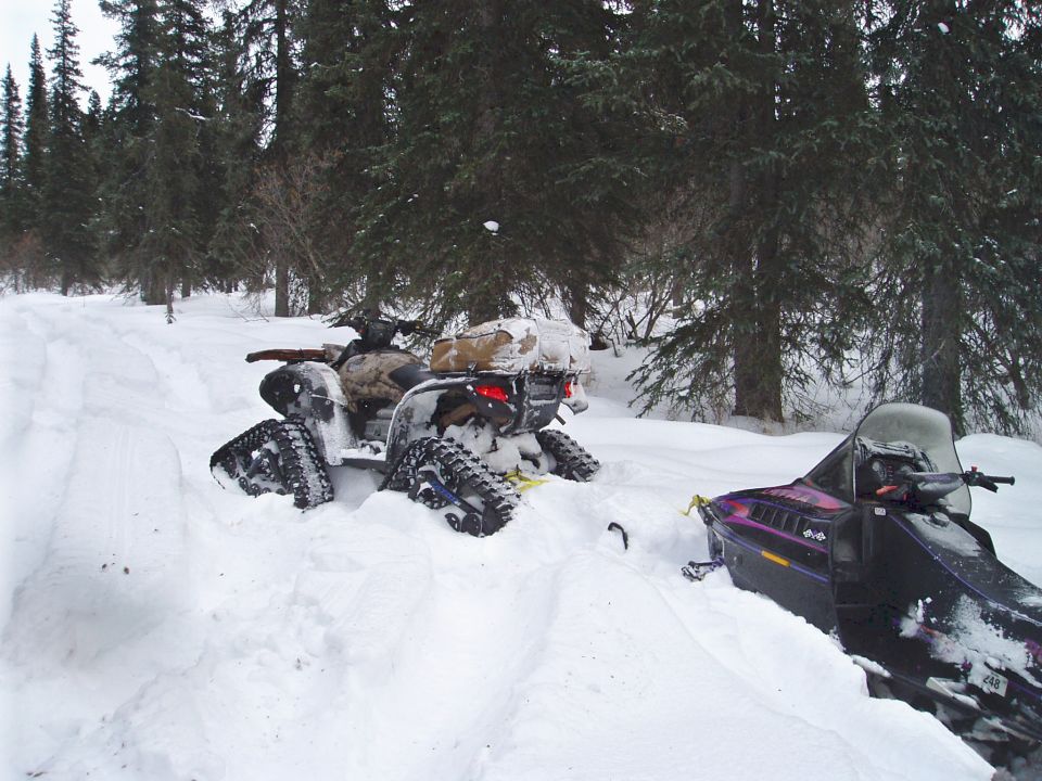 ATV with tracks stuck in snow next to a snowmobile in a forest