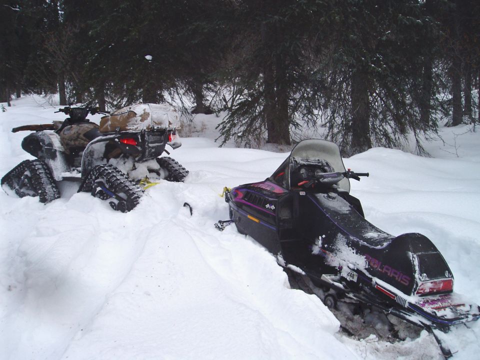 Two snowmobiles and an ATV stuck in deep snow with trees in the background