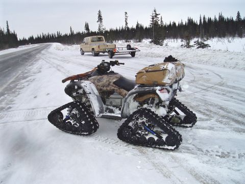 2000 Ford E350 - An ATV equipped with tracks sits on a snow-covered road beside a pickup truck towing a trailer.