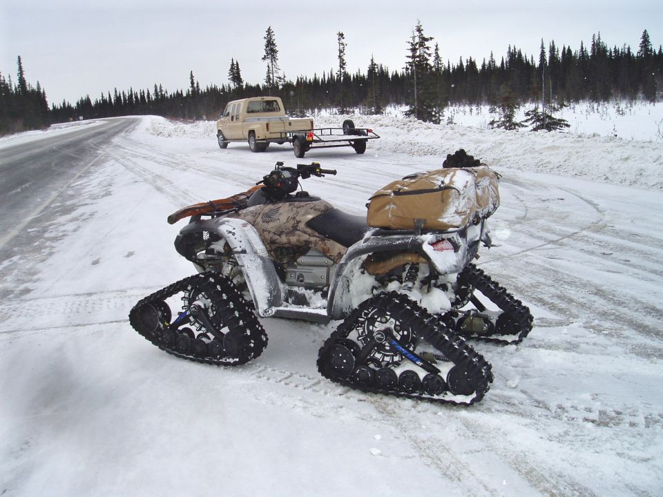 ATV with tracks in snow, parked next to a pickup truck and trailer on a snowy road