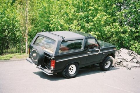 1985 Ford Bronco - This black Ford Bronco is parked outdoors near a pile of debris and lush green trees.
