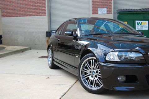 2002 BMW M3 - A black BMW M3 coupe is parked in an industrial area with a brick building and dumpsters in the background.