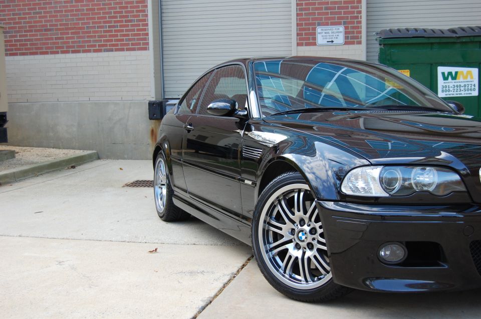 Black BMW M3 coupe, front 3/4 view, parked on concrete near brick building and dumpsters