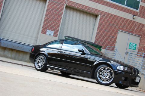 2002 BMW M3 - A black BMW M3 is parked in front of a brick building with large roll-up doors.