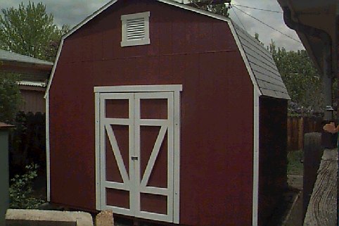 Dark red gambrel-roof storage shed with white double doors in a backyard.