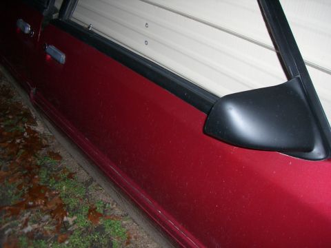 1988 Ford Bronco - Side view of a red vehicle door and exterior mirror, parked near a garage.