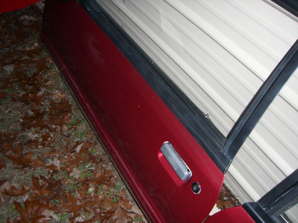 Red car door with chrome handle and lock cylinder, leaning against a white corrugated wall. Fallen leaves on the ground