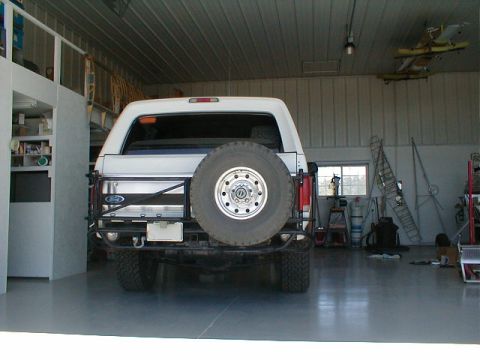 1984 Ford Bronco - Rear view of a white Ford Bronco with a spare tire and custom bumper inside a garage.