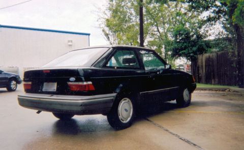 1989 Ford Escort - This black sedan is parked on a wet surface next to a building.