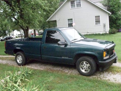 1995 GMC Sierra 1500 - A dark green 1995 GMC Sierra 1500 pickup truck is parked on a gravel driveway in front of a house.