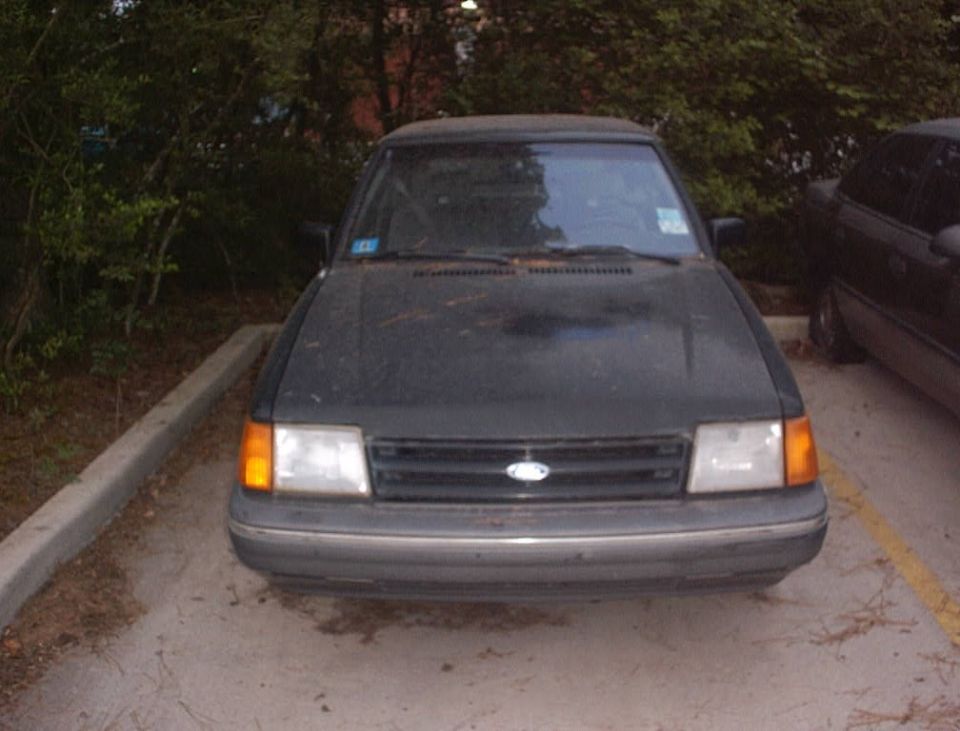 Dirty dark gray Ford Escort parked in a lot, front view showing grille and amber corner lights