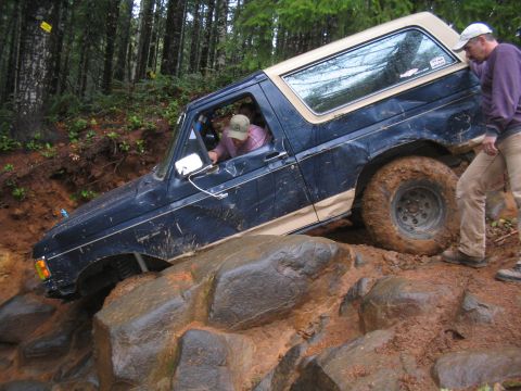1989 Ford Bronco - A Ford Bronco maneuvers over large rocks and mud on an off-road trail, with two people inside and beside the vehicle.